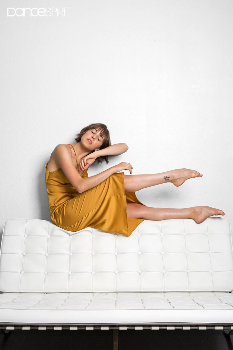 Gianna Reisan sat with her head facing towards the camera, her eyes closed, and leaning against her left arm as if sleeping. Her legs are pointed to the right of the photo, her right leg above the left, but both parallel to each other.