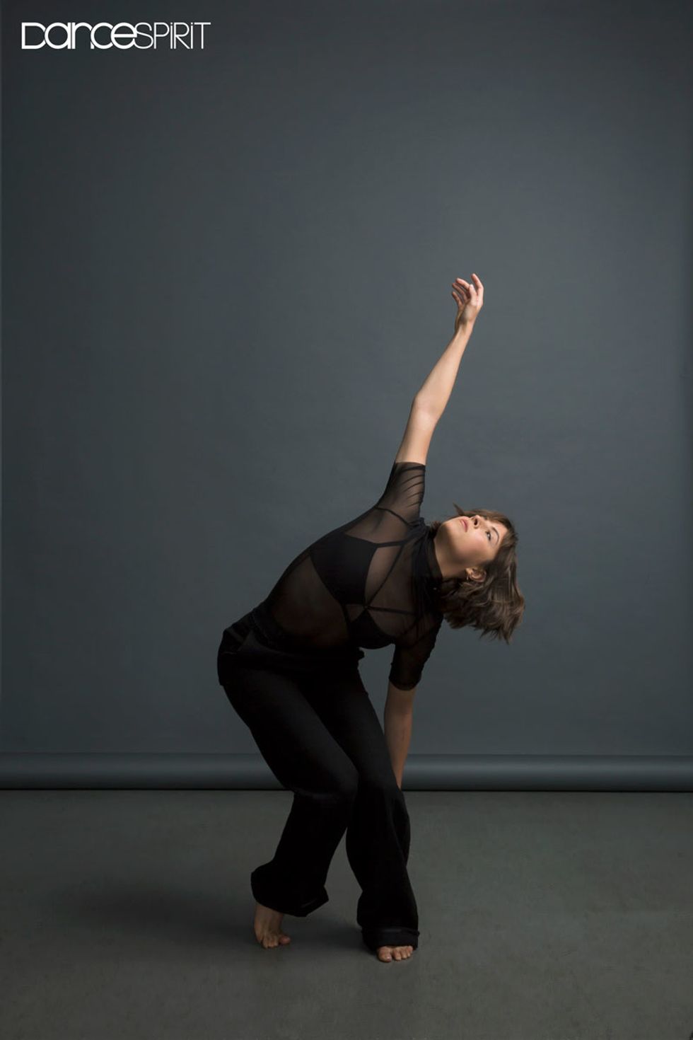 Gianna Reisan with her right foot slightly in front of the left. Her left arm is pushed down towards her left foot, whereas her right arm is outstretched to the ceiling above her. She is facing upwards.