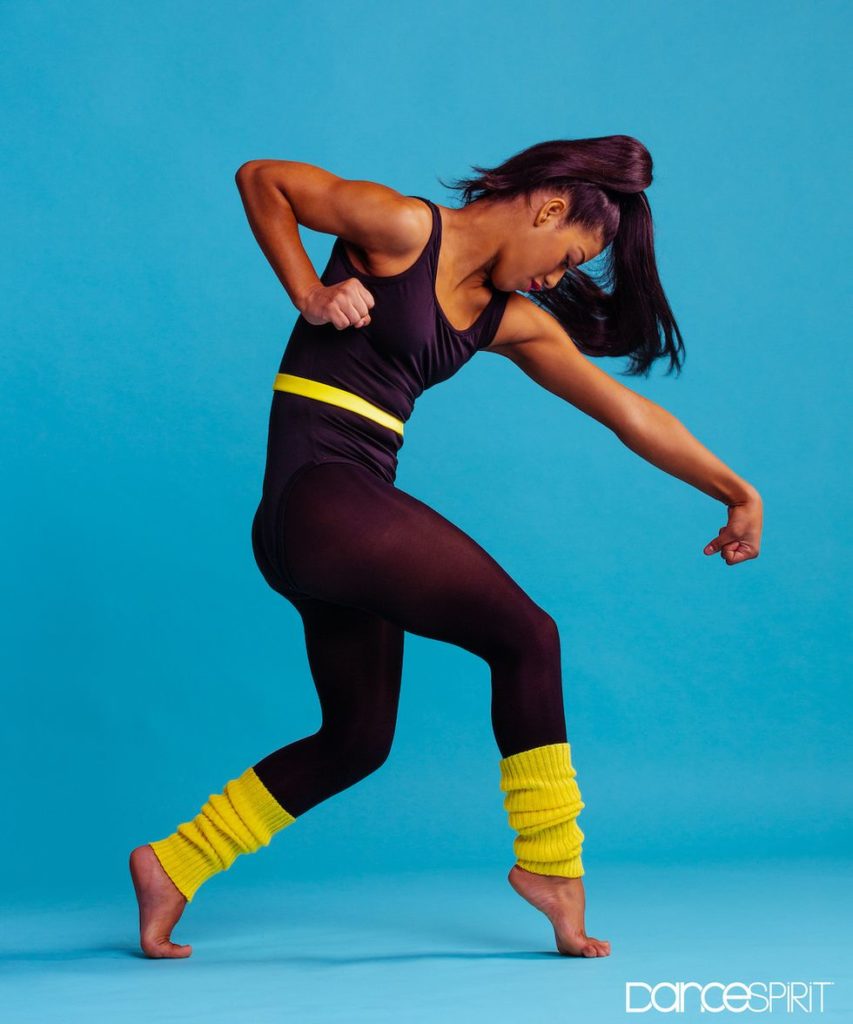 15-year-old Selena Hamilton, wearing a black leotard and neon yellow legwarmers, she is standing on pointed toes, her head is tilted downwards and her right arm is pulled back and her left arm is stretched out.