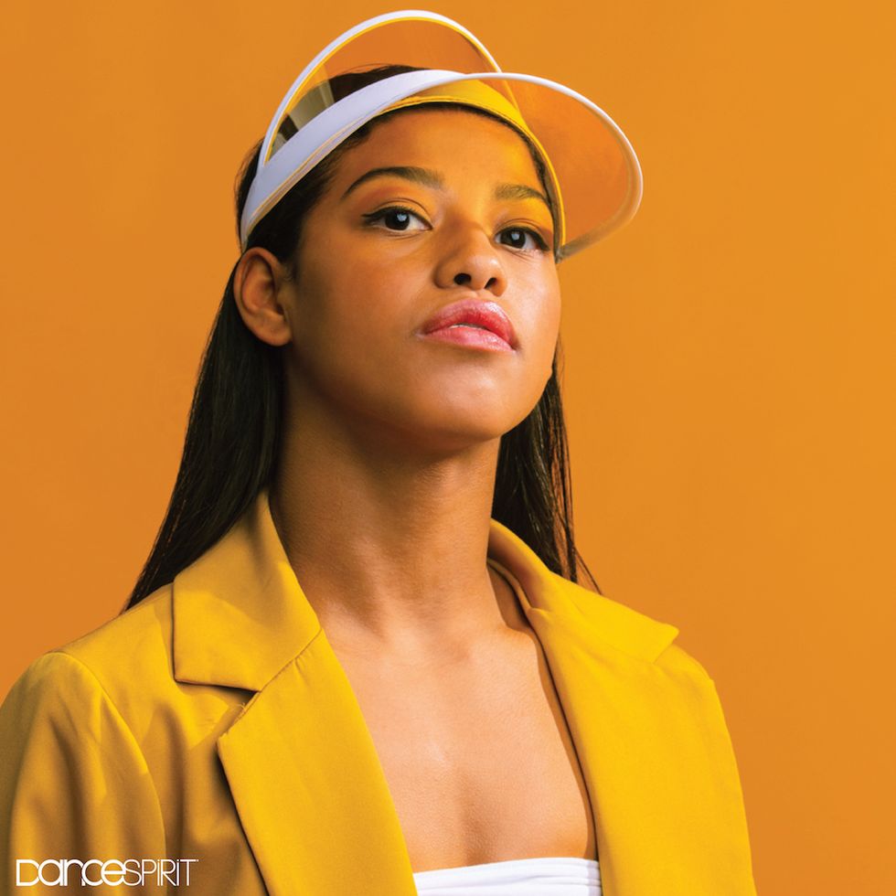 A close up photo of Selena Hamilton wearing a bold yellow blazer and white and yellow visor. She is facing towards the camera, with her chin held high.