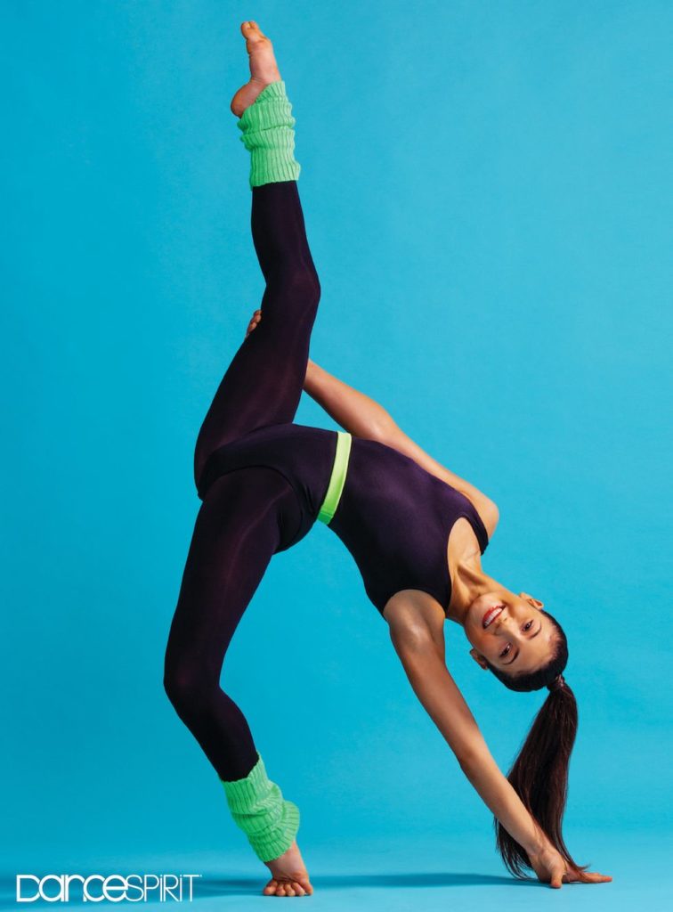 Dyllan Blackburn, wearing a black leotard and neon green legwarmers, pulling her right leg skywards and facing the camera against a blue background, her head pushed downwards so it is close to her left supporting arm.