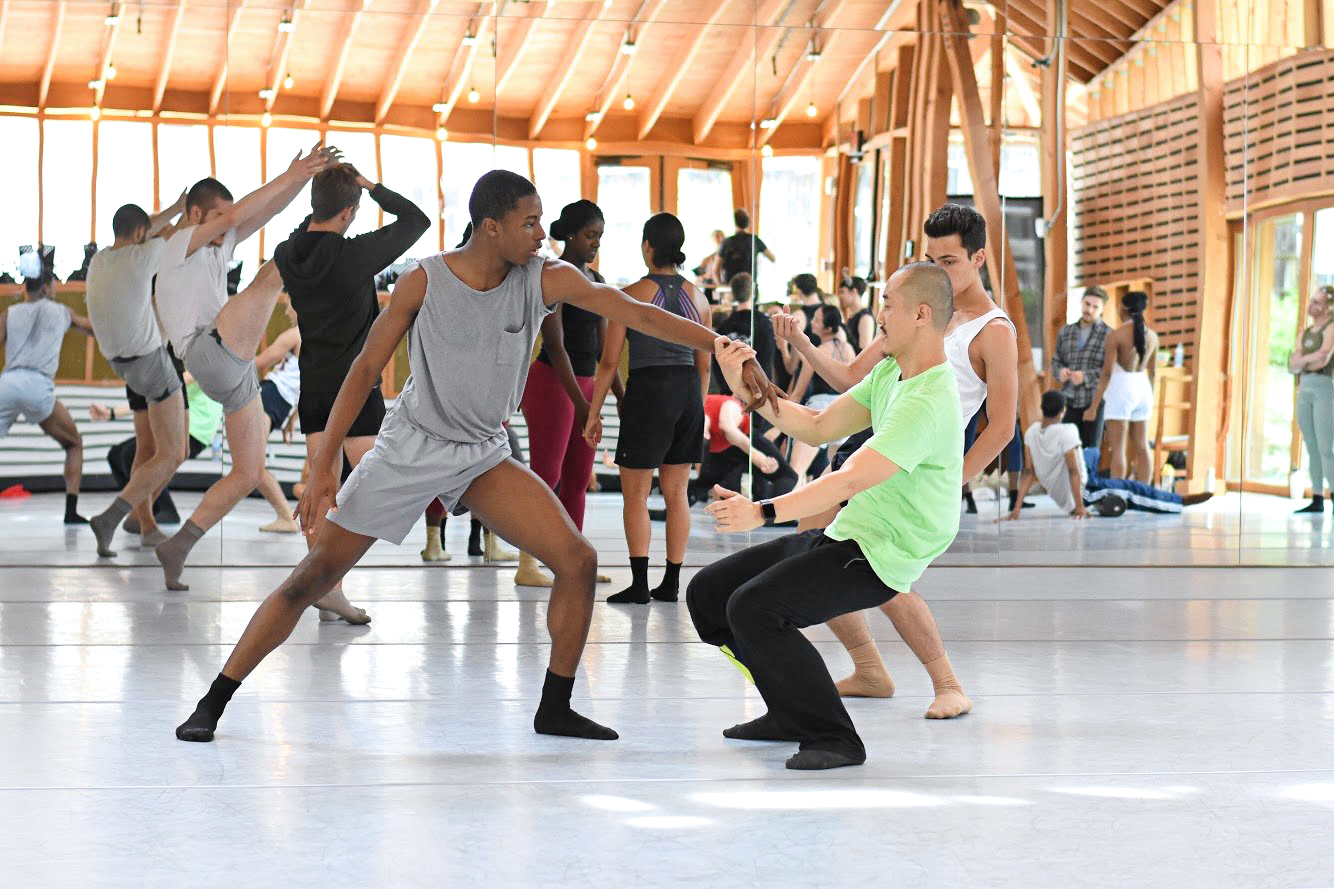 Jamaii Melvin stands in a lunge while his dance partner hinges, holding onto his left arm. Jamaii is wearing a gray top and gray shorts.