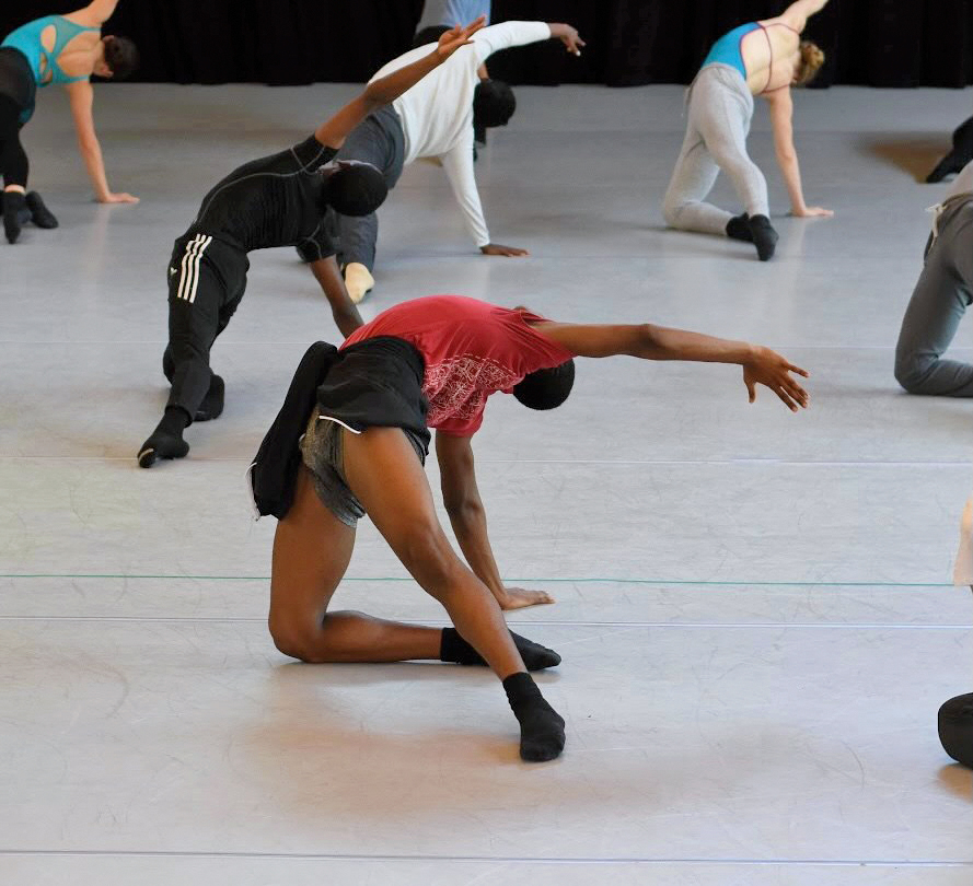 Jamaii Melvin kneels on the floor with his back arched and his head and left arm reaching toward the sky. 3 pre-pro dancers share the dancewear they live in.