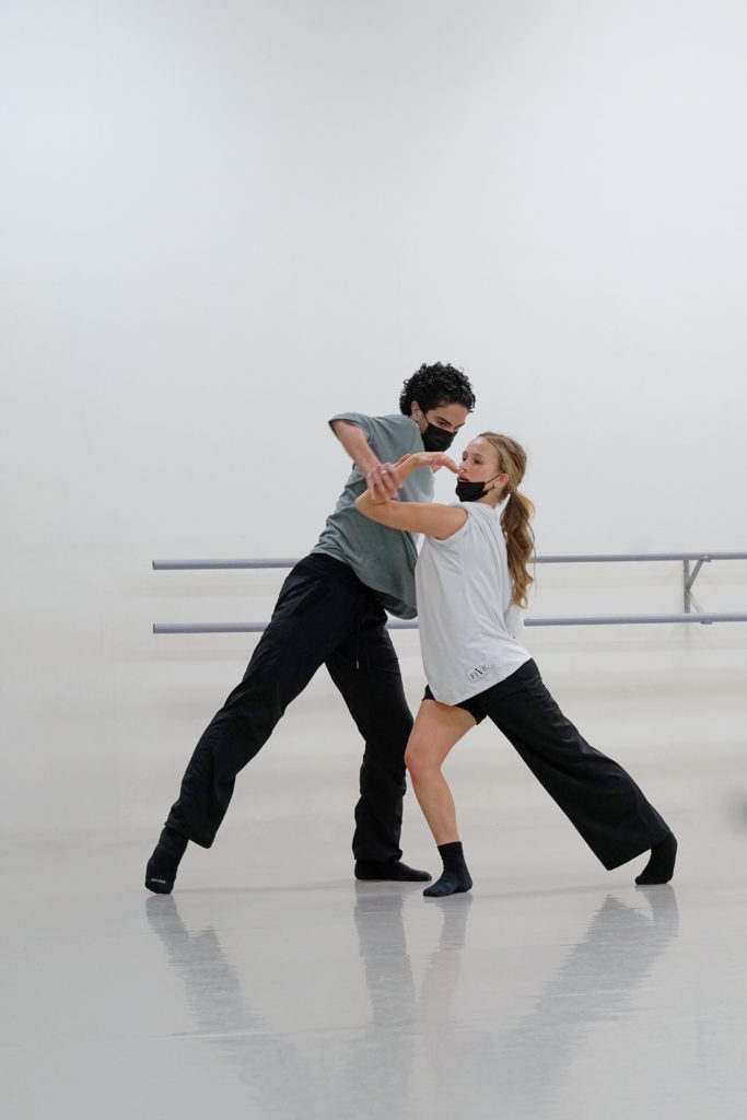 Ella Horan and her dance partner do contact improv. They are lunging toward each other and he has his hand pushing down on her left arm. She is wearing a white shirt and long pants with one leg bunched up. 3 pre-pro dancers share the dancewear they live in.