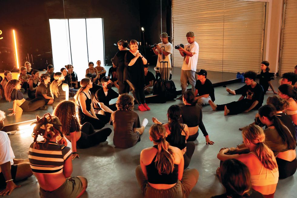 Leaning against a studio mirror, choreographer Tessandra Chavez smiles as several students sit on the floor in front of her.
