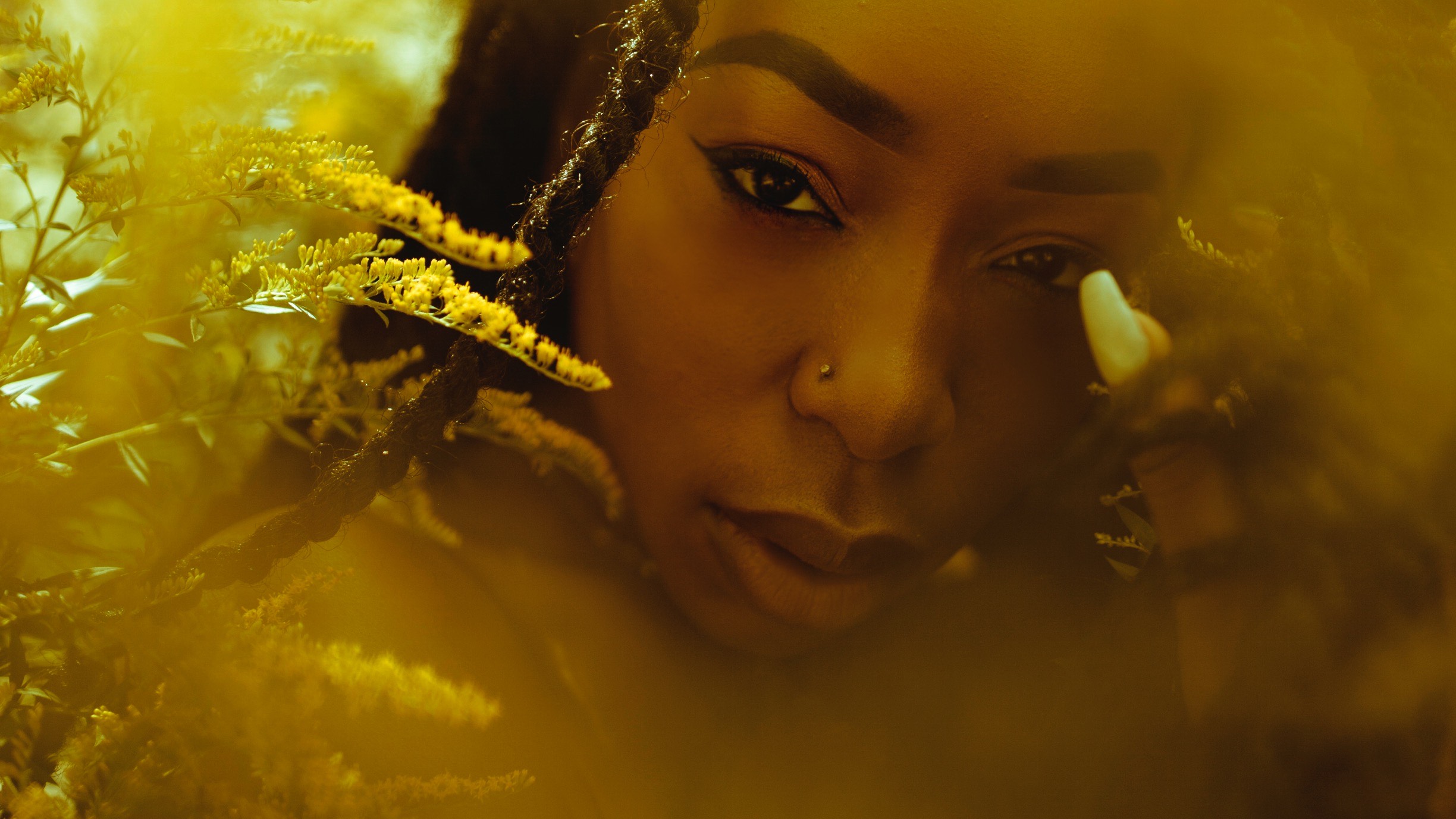 A close up shot of Amanda, a Black woman gazes into the lens through some plants.