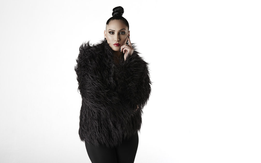 Wearing a black fur coat, Tricia poses with one hand against her cheek.