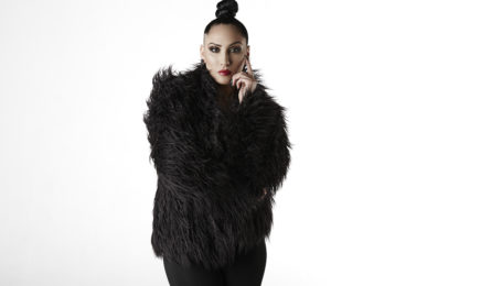 Wearing a black fur coat, Tricia poses with one hand against her cheek.