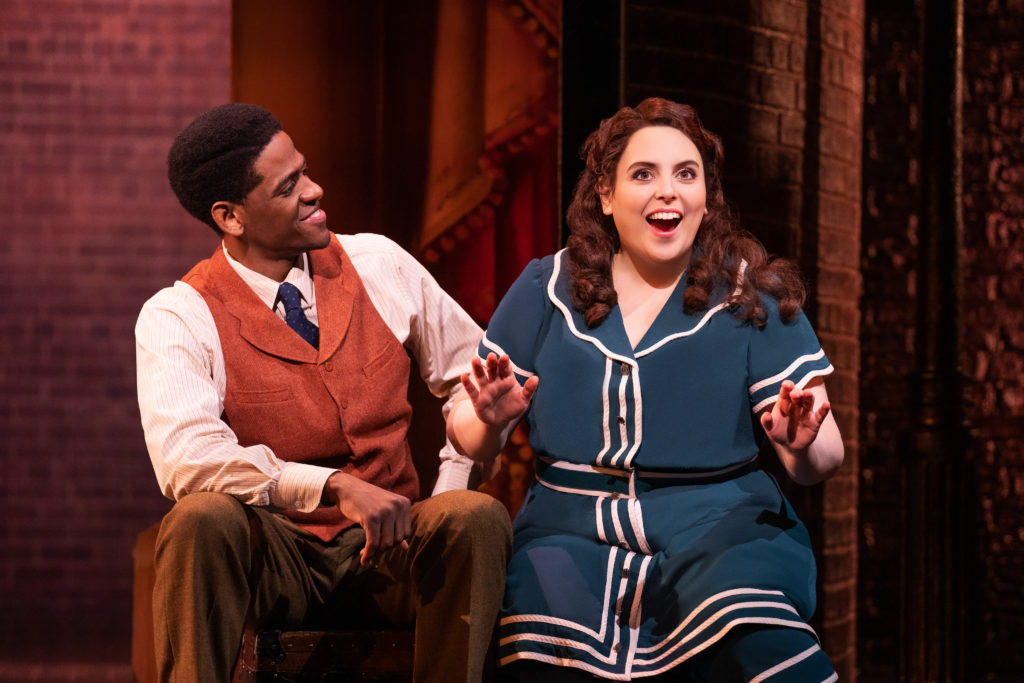 Jared Grimes (as Eddie Ryan), wearing a brown suit, white shirt and blue tie, looks at Beanie Feldstein (as Fanny Brice), who is seated on his left. She is wearing a blue and white dress. Feldstein is looking enthusiastically ahead, hands stretched in front of her.