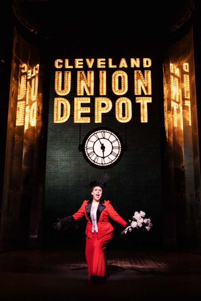 Beanie Feldstein as Fanny Brice, wearing a red and black skirt suit, under a brightly lit sign that reads, "Cleveland Union Depot."
