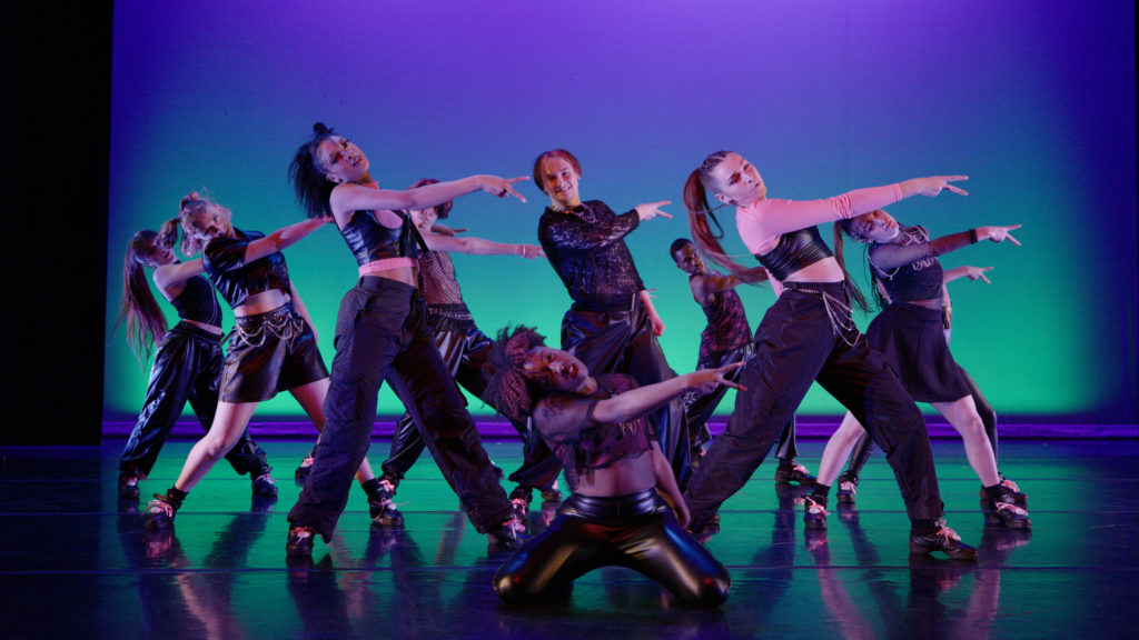 A group of dancers in black shoes and clothing do a synchronized dance in a tight formation.