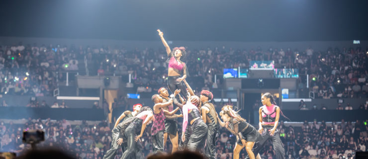 A group of dancers performing for a full stadium at L.A. KCON. One dancer is being held in the air by the others.