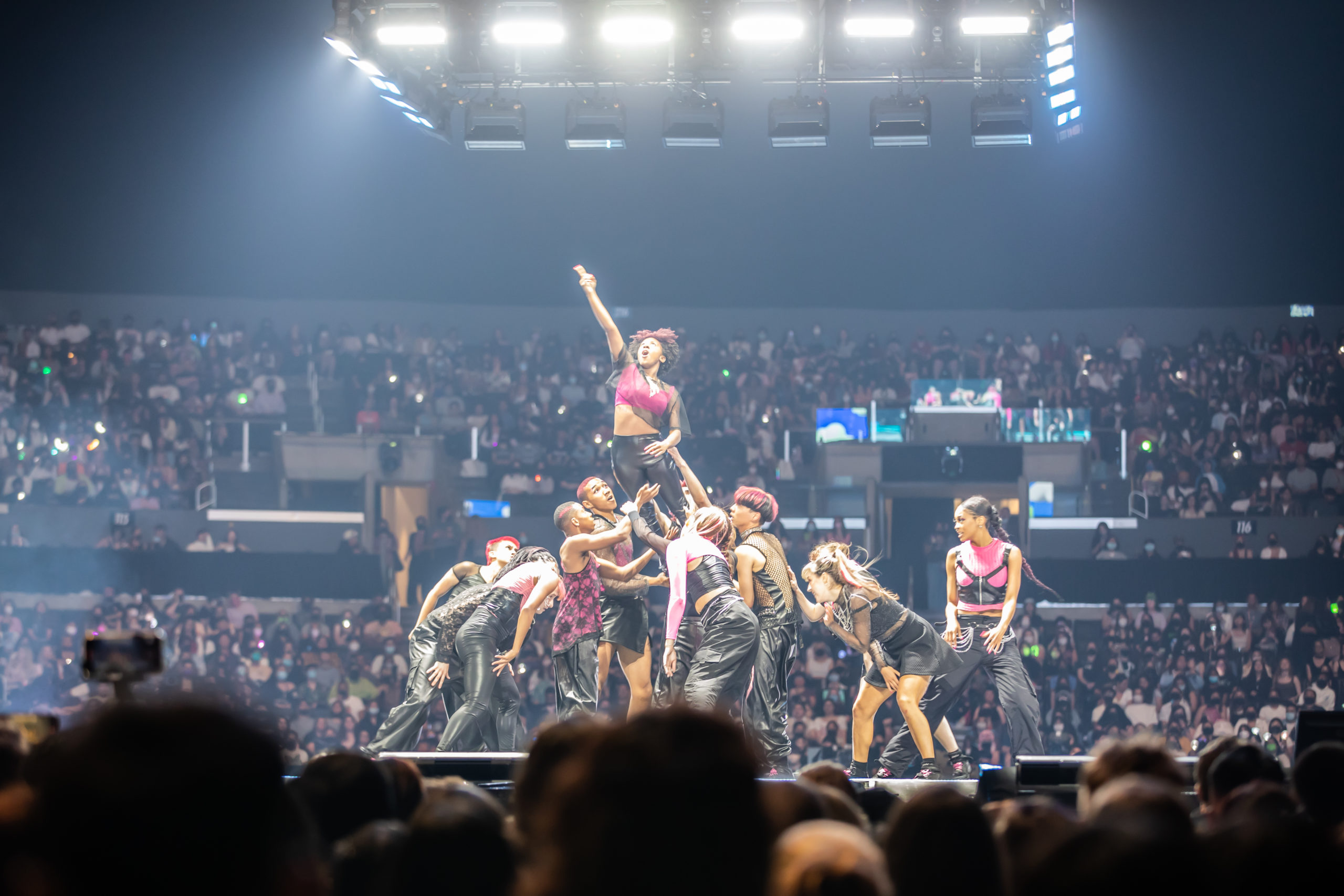 A group of dancers performing for a full stadium at L.A. KCON. One dancer is being held in the air by the others.