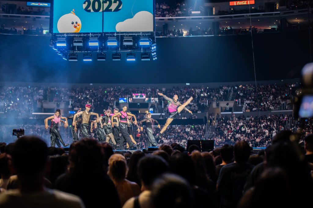 A group of dancers performing for a full stadium at L.A. KCON. One is doing a split leap.