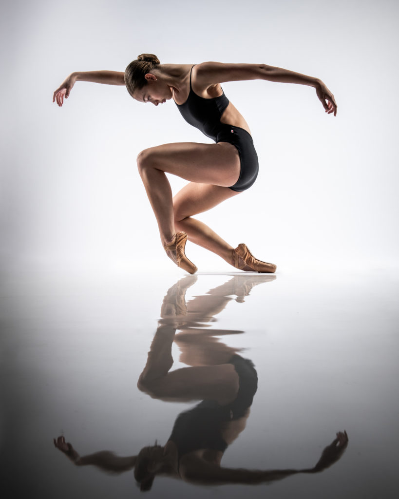 A ballet dancers in pointe shoes sinks toward the floor, her shape reflected in the glossy, white floor.