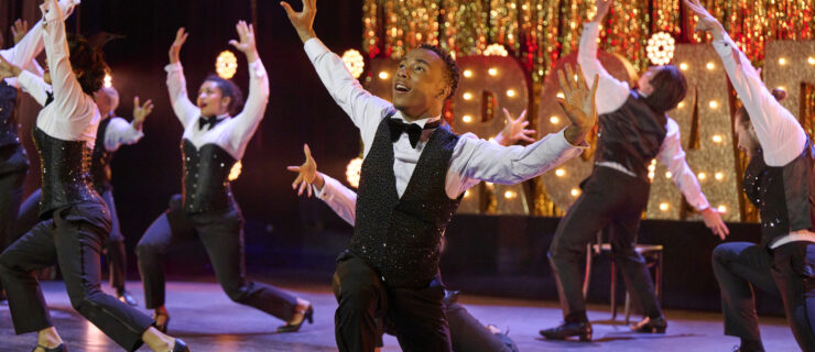 Against a glittery gold backdrop, the 8 remaining "SYTYCD" competitors, wearing glammed-up tuxedos, are caught mid-performance, their arms raised, hopeful expressions on their faces.