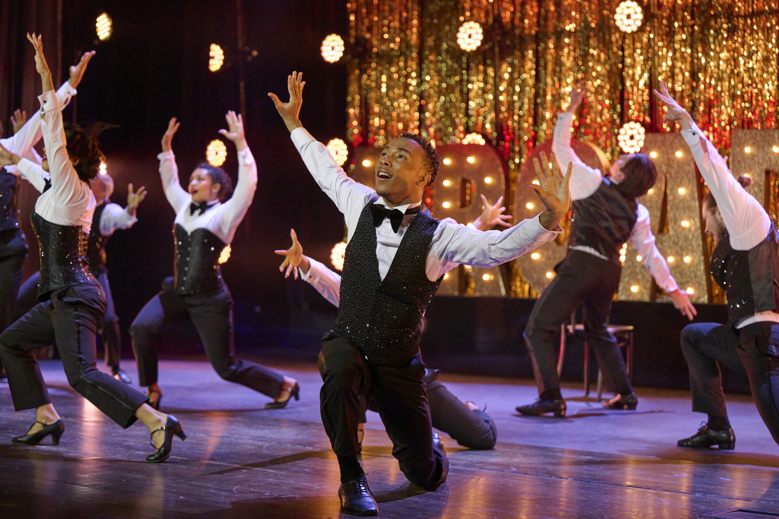 Against a glittery gold backdrop, the 8 remaining "SYTYCD" competitors, wearing glammed-up tuxedos, are caught mid-performance, their arms raised, hopeful expressions on their faces.
