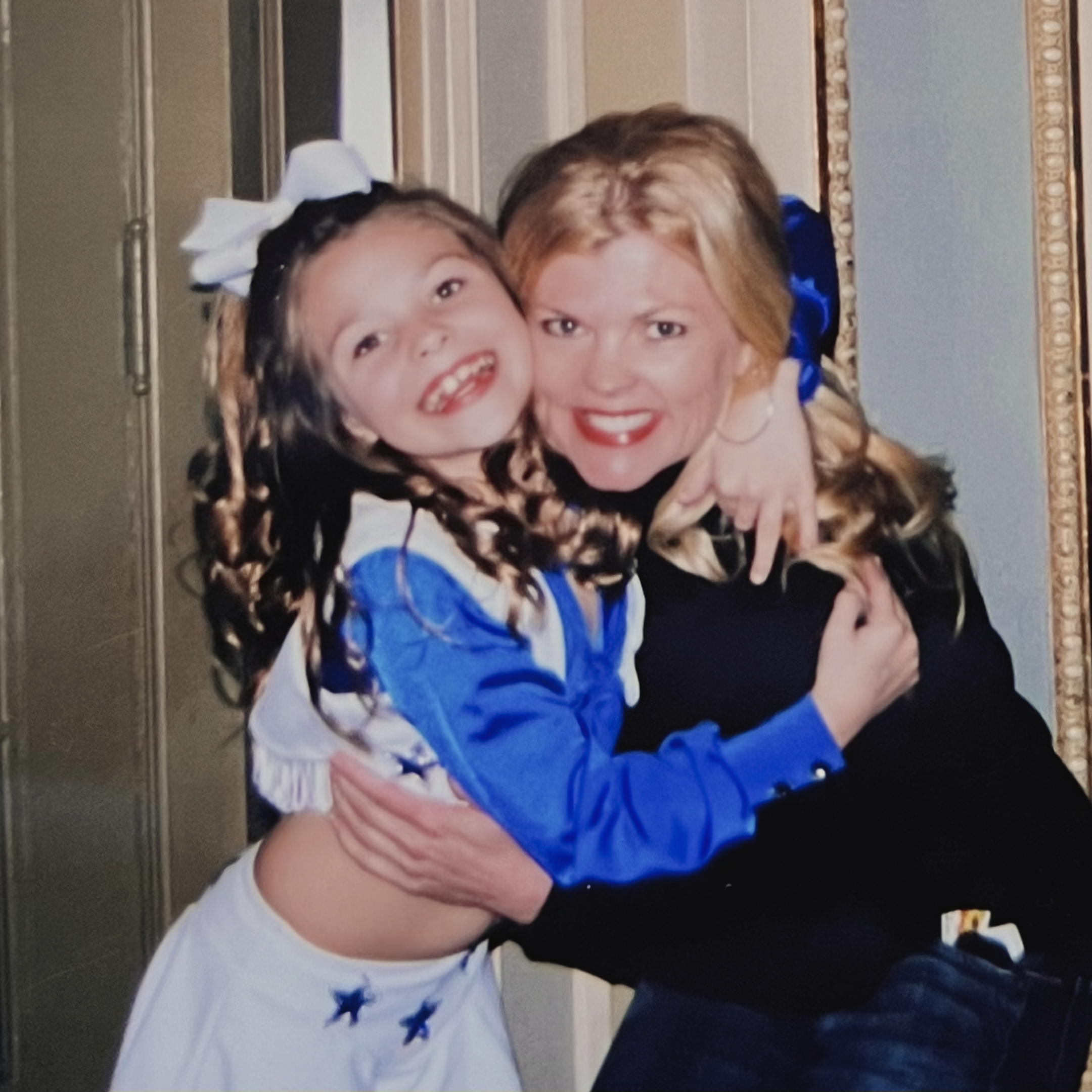 A blonde woman hugs a young girl wearing a blue and white cheerleading uniform 