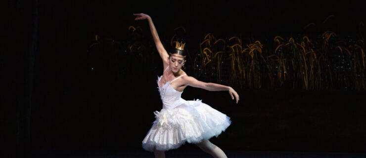 a female dancer wearing a white tutu as she lunges on stage with her arms extended