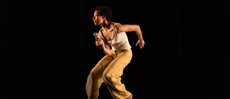 Eleni Loving lunging while holding a microphone on stage. The lighting is dark and she is wearing a tank top with yellow pants