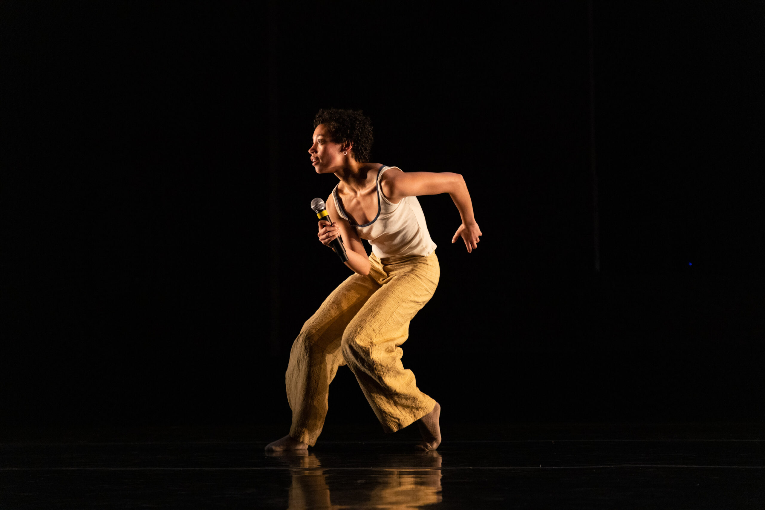 Eleni Loving lunging while holding a microphone on stage. The lighting is dark and she is wearing a tank top with yellow pants