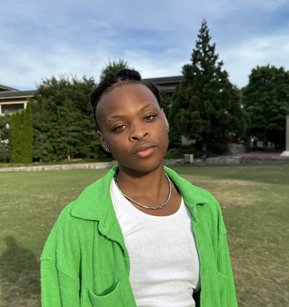 Ilo E's headshot outdoors wearing a green jacket and white tank 