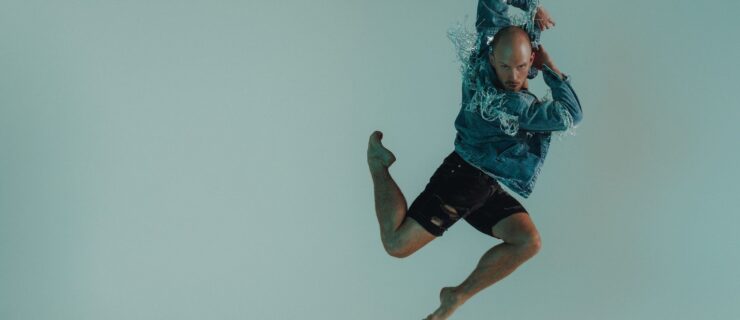 a male dancer jumping in the air in a stag position with his arms swinging back. He's wearing a blue jacket and black shorts