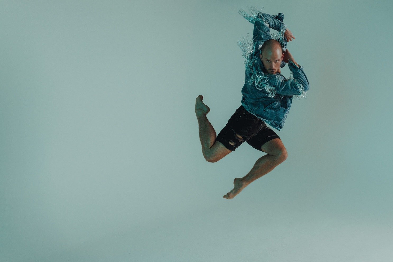 a male dancer jumping in the air in a stag position with his arms swinging back. He's wearing a blue jacket and black shorts