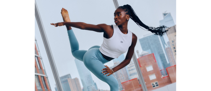 Caroline T Dartey wearing blue leggings and a white crop top dancing in a studio with large windows overlooking the city