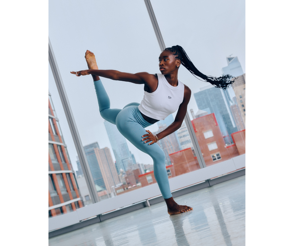 Caroline T Dartey wearing blue leggings and a white crop top dancing in a studio with large windows overlooking the city