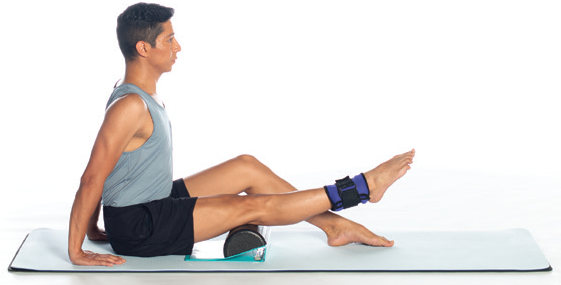 A dancer sitting with his legs propped on a foam roller. He has an ankle weight on his right leg and is lifting the foot off the floor.