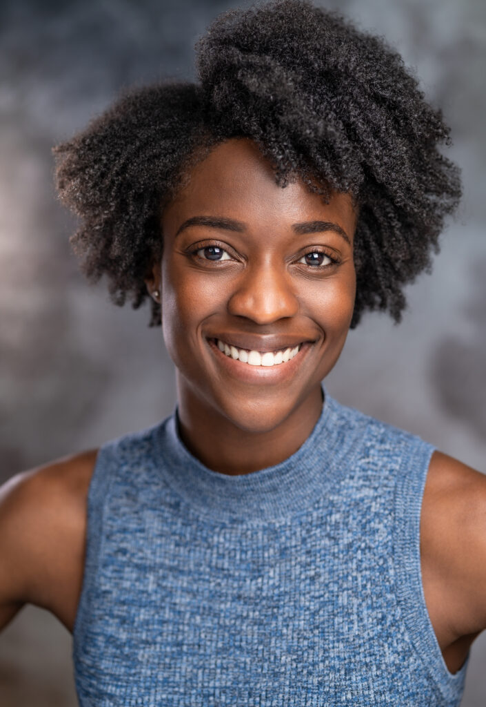 Crystal Nicholls. She wears a blue tank top while standing against a grey backdrop 