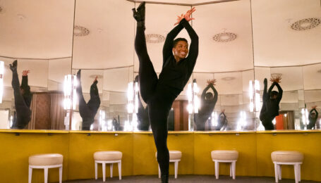 Karmen Moore kicking her leg into a tilt with her arms over her head. She dances in a dressing room with surrounding mirrors