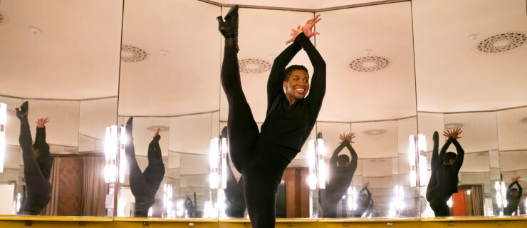 Karmen Moore kicking her leg into a tilt with her arms over her head. She dances in a dressing room with surrounding mirrors