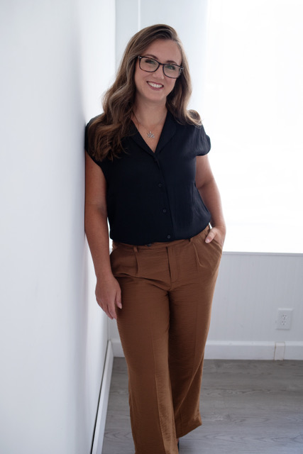 Kristen Mackel's headshot. A woman wearing brown pants and a black top smiling while leaning against a white wall 