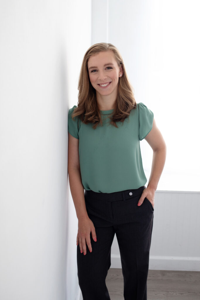 Leigh Skvarla's headshot. A woman wearing jeans and a green top smiling while leaning against a white wall 