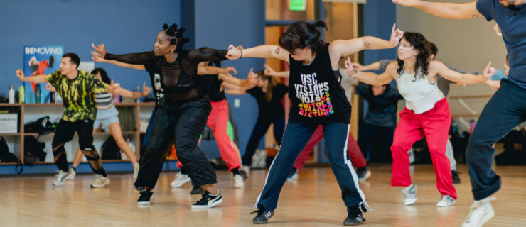 Hip hop dancers in a studio dancing together as a group. They point their hands out to either side and tap their right foot.