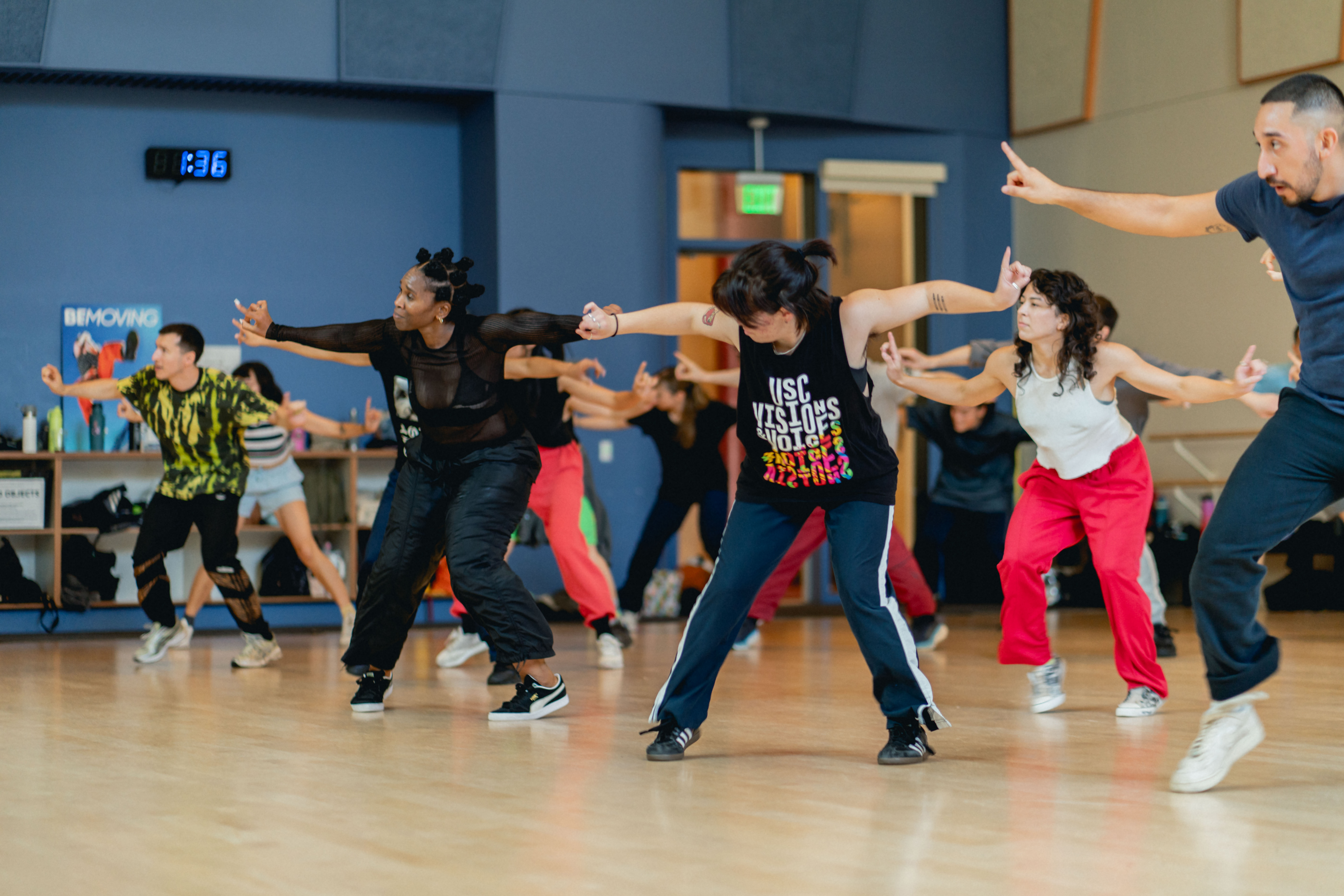 Hip hop dancers in a studio dancing together as a group. They point their hands out to either side and tap their right foot.