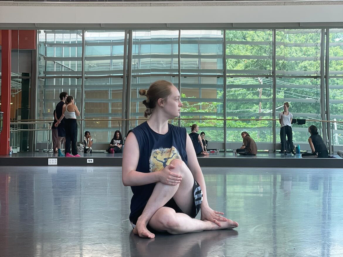 Aubrey Mailloux sits on the studio floor, stretching with one leg crossed over the other