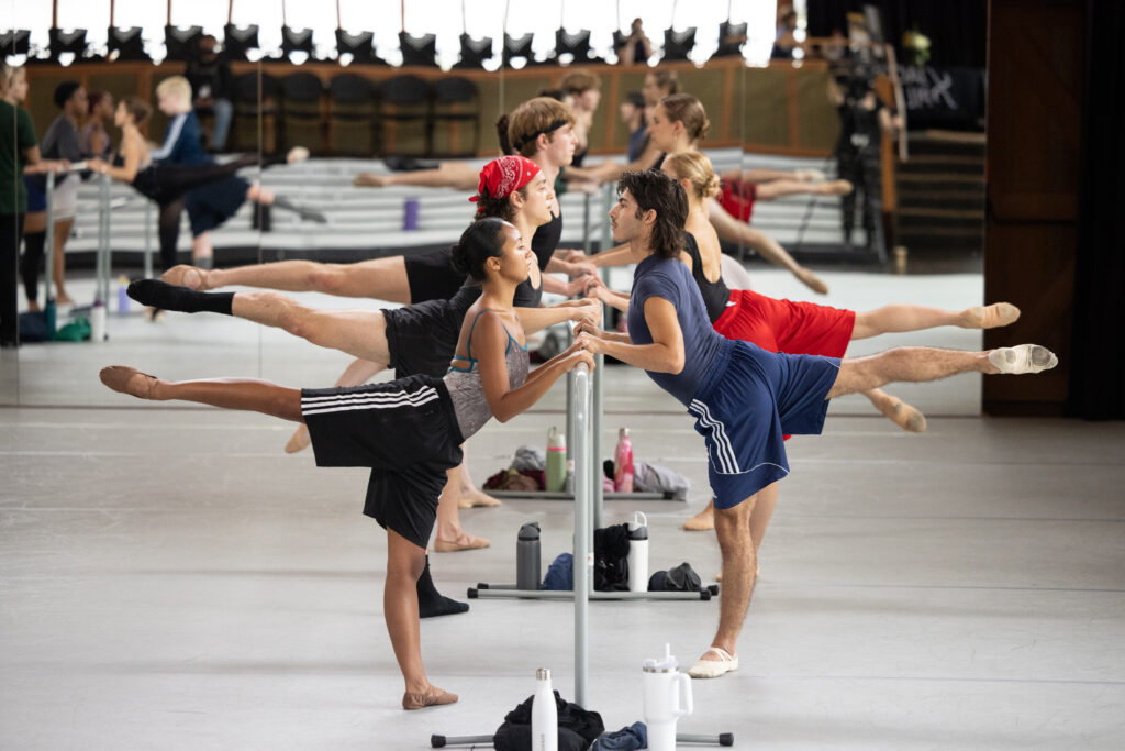 Dancer in large studio facing handrail while raising right and left in arabesque 