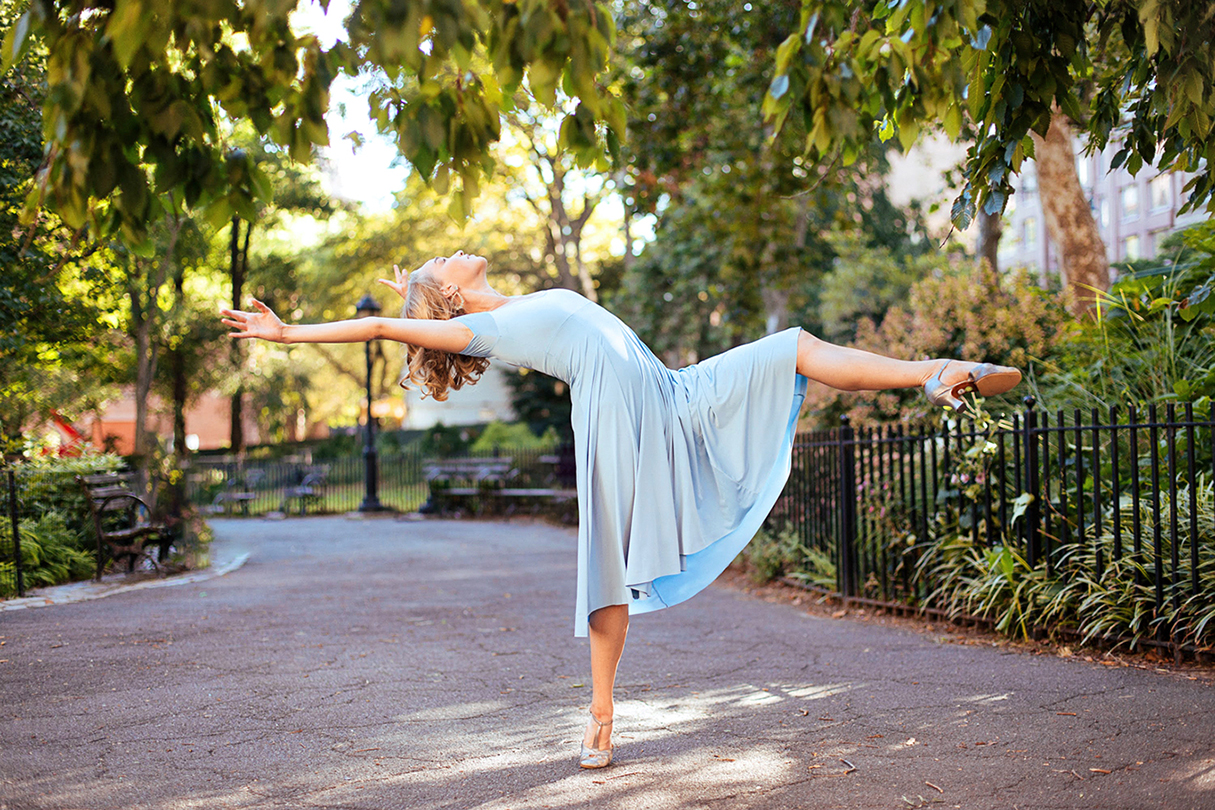 Kathleen Laituri dancing outside in a park. She wears a long blue dress and character heels as she executes a layout.