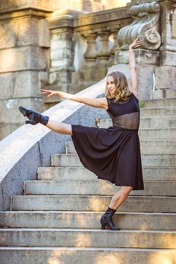 Kathleen Laituri dancing on stone steps. She wears a black dress and dance boots while extending her leg a la seconde 