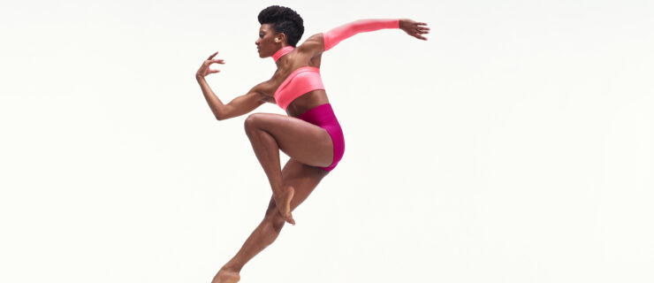 Jacquelin Harris wearing an asymmetrical two-toned pink leotard while jumping in the air against a white backdrop