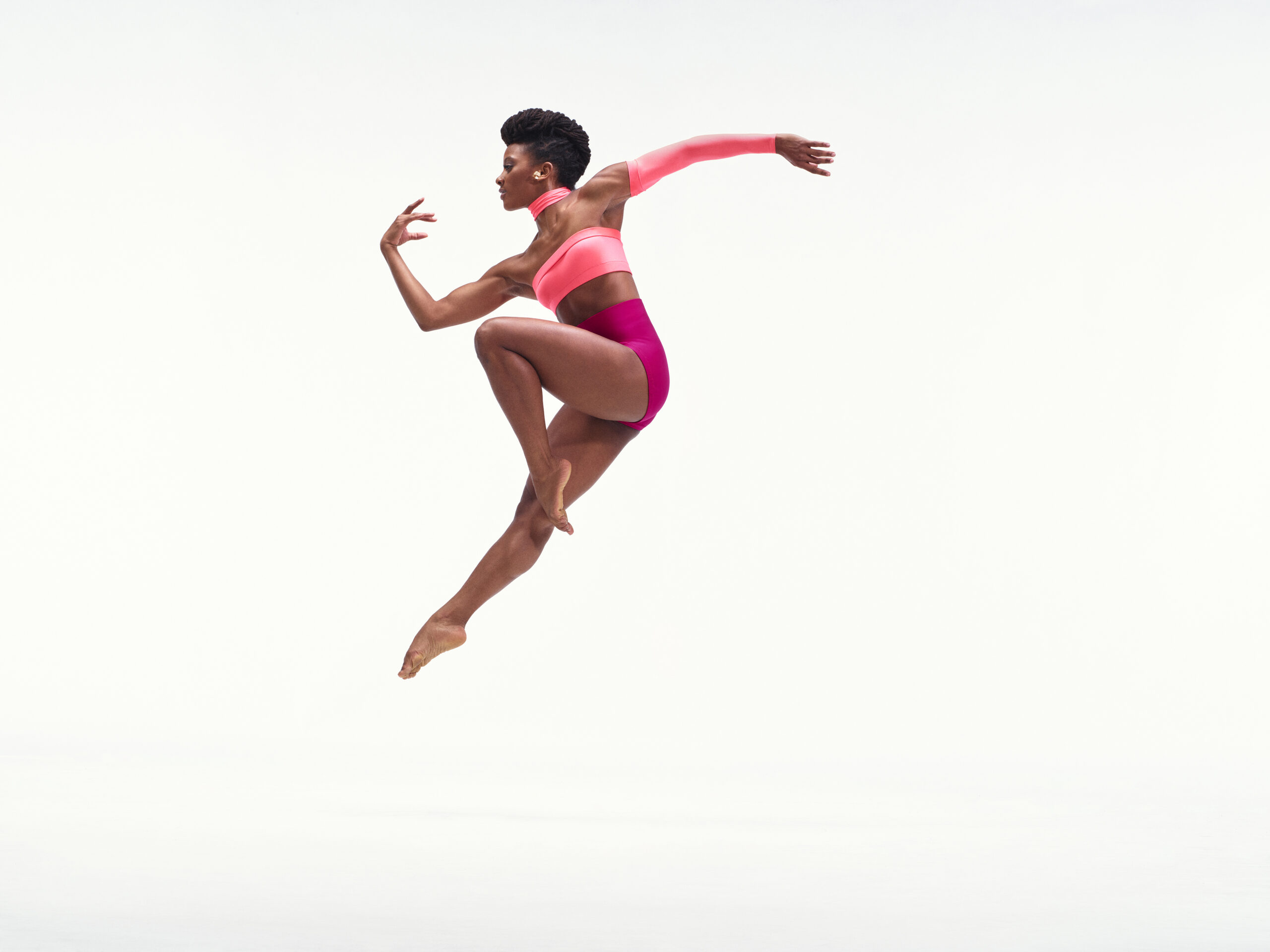 Jacquelin Harris wearing an asymmetrical two-toned pink leotard while jumping in the air against a white backdrop