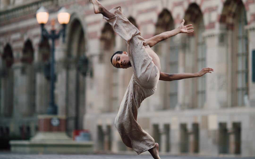 Braylon Browner lifting his leg over his head with his arms swinging behind him. He wears flowy tan pants and dances in front of a brick building.