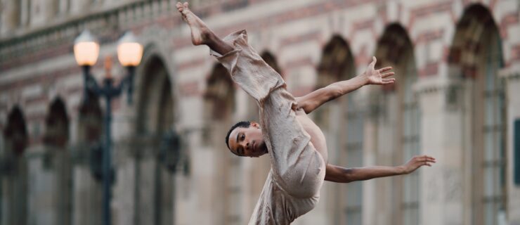 Braylon Browner lifting his leg over his head with his arms swinging behind him. He wears flowy tan pants and dances in front of a brick building.