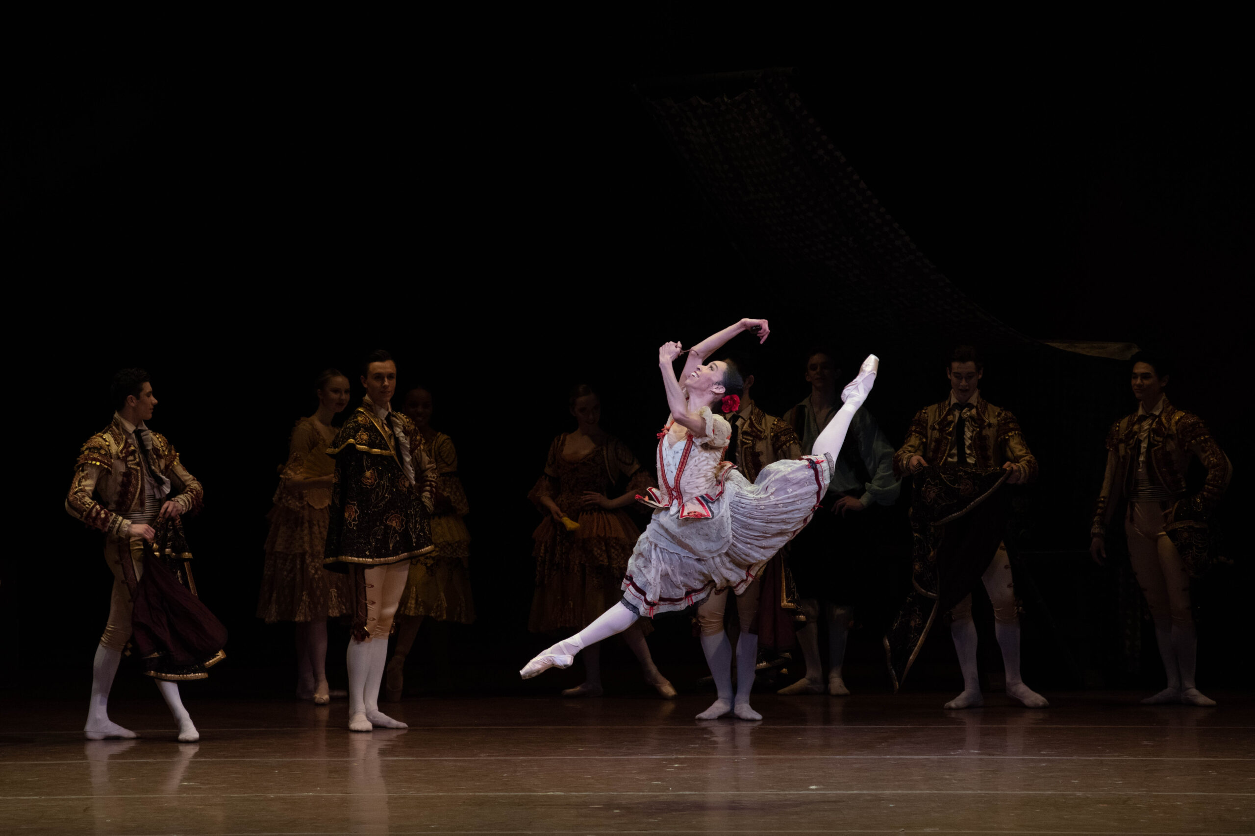 A female ballerina performing a firebird jump onstage. She wears a peasant style dress and pointe shoes.