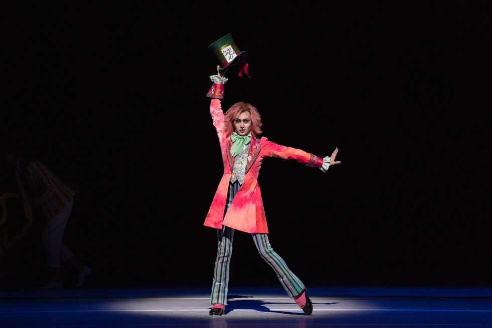 Against a stark black background and in a spotlight, Amelia Townsend poses as the Mad Hatter in a red coat with a spiky red wig and stark makeup.
