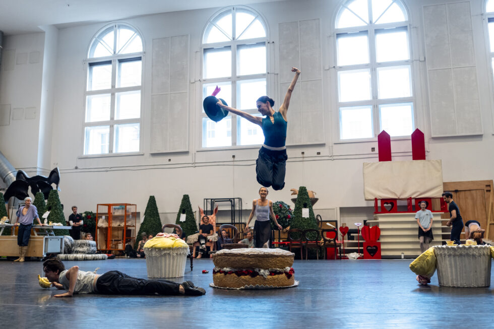In rehearsal, Amelia Townsend as the Mad Hatter jumps off a cake-shaped set piece, her feet tucked behind her body and her arms in the air.