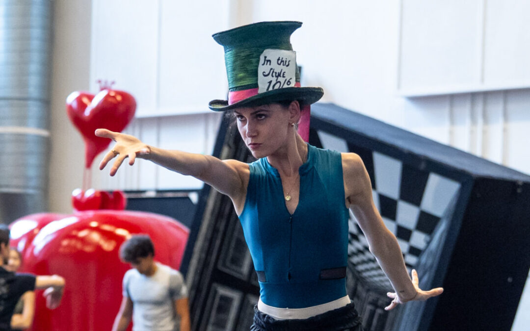 In a blue leotard, black pants, and the Mad Hatter's oversized top hat, Amelia Townsend points dramatically at something off camera.